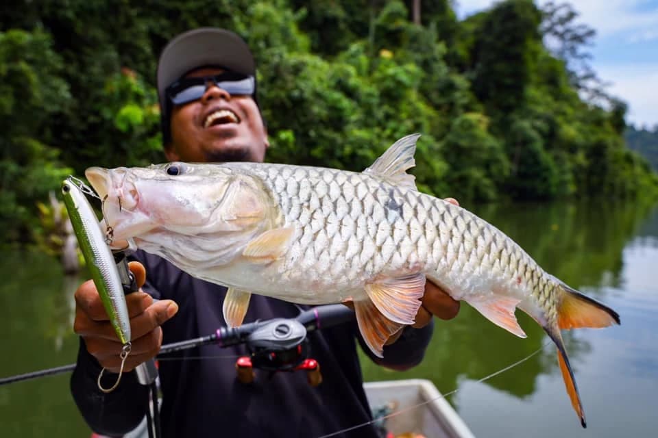 An angler sharing a freshwater catch beside a tree-lined lake.
