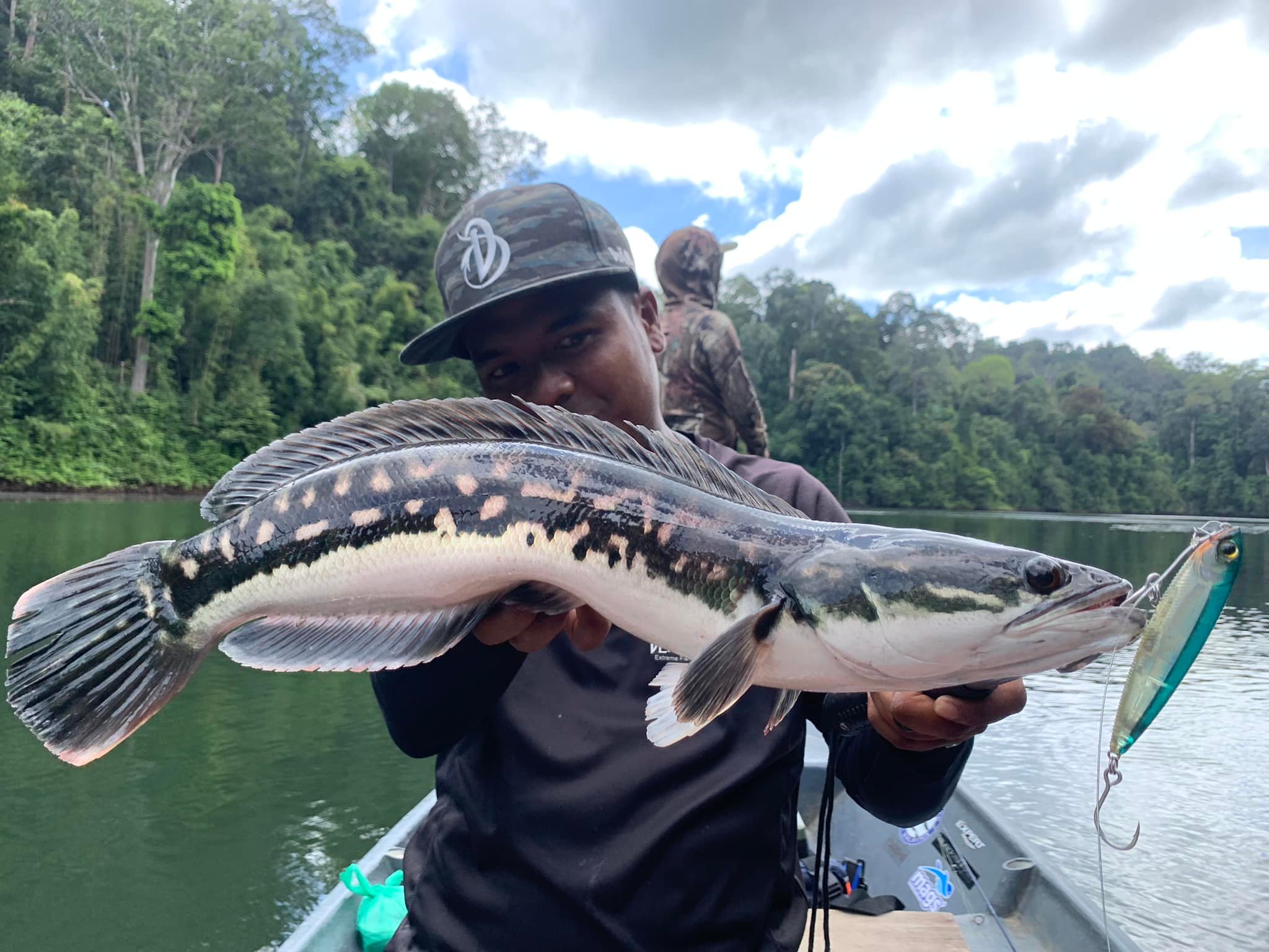 An angler holding a toman with forest and freshwater behind him